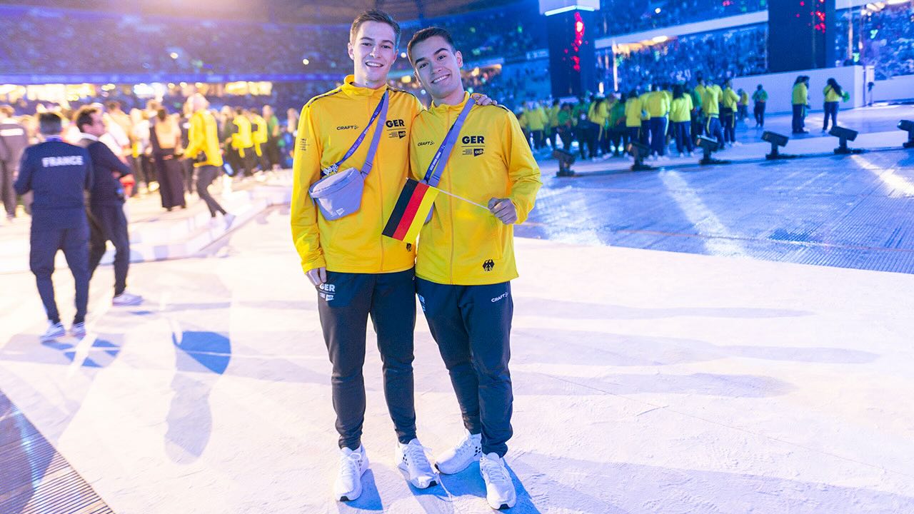 Elia Pörsch steht links und Jona Pörsch steht rechts, beide haben jeweils einen Arm um den anderen gelegt. Sie tragen beide gelbe Jacken mit dem Aufdruck GER und dem Bundesadler und schwarze Trainingshosen mit dem Logo GER. Jona Pörsch hält eine kleine Deutschland-Fahne in seiner linken Hand. Im Hintergrund ist ein belebter Austragungsort der Rhine-Ruhr 2025 FISU World University Games mit Besucherrängen zu erkennen.