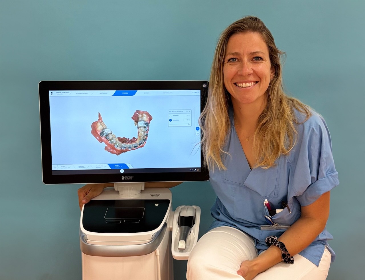 Die Preisträgerin Dr. Eva May Schraml sitzt rechts neben einem fahrbaren Computer mit Intraoralscanner mit der Darstellung eines Intraoralscans auf dem Bildschirm und lächelt in die Kamera.