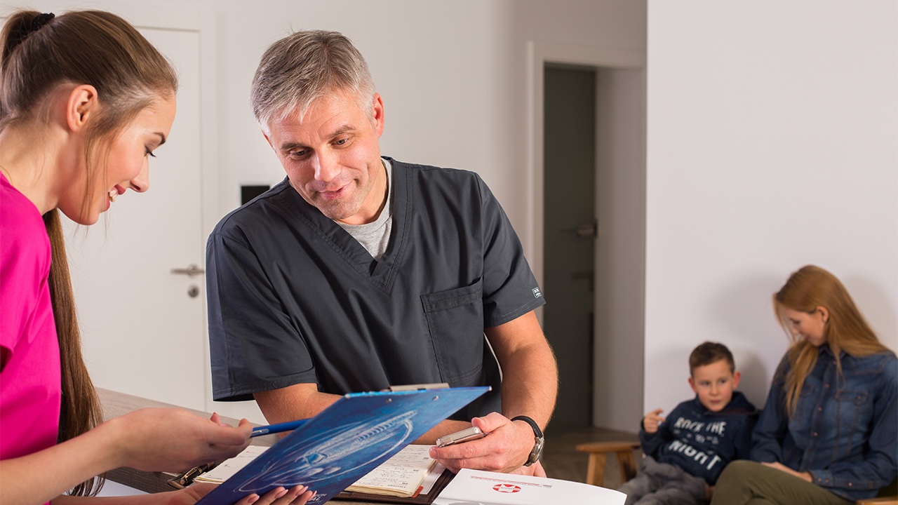 Gesprächssituation an einer Praxistheke, links eine Fraumit rosa Shirt, in der Mitte ein Mann mit dunkelgrauem Praxiskittel, im Hintergrund wartende Patienten.